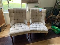 Pair of beige tufted side chairs with wooden legs positioned on hardwood floor near window, well-lit indoor setting.