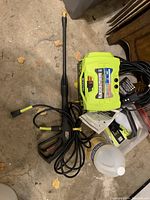 Photo showing the Ryobi pressure washer unit with connected high-pressure hose, spray gun handle, wand, power cord and partial jug of cleaner on floor.