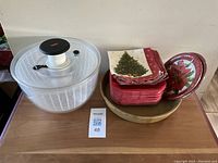 Photo of white plastic lettuce spinner with clear lid and black knob, Christmas-themed small square plates and napkins stacked, round wooden plate/board underneath.