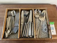 Wooden flatware organizer with multiple compartments containing polished stainless steel forks, spoons, knives, and serving utensils with ribbed handles.