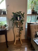 Front view of tall white decorative plant stand with Pothos plant on top near windows and upholstered chair.