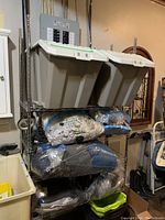 Full view of the steel shelving unit with two large plastic bins on top and several sealed plastic bags containing folded textiles on the shelves below.