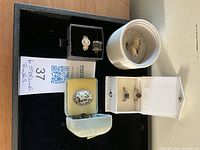 Photo showing five sterling rings in different display boxes and a white round container, placed on a black surface with handwritten label.