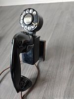 Black rotary dial antique wall phone with cloth-covered cord shown from an angled side view highlighting handset and dial.