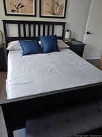 View of black IKEA Hemnes queen bed frame with white mattress and two blue pillows. Headboard has vertical slats and solid footboard is visible. Nightstand with two drawers and round knobs to right of bed.
