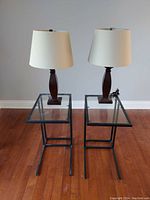 Two IKEA Vittsjo glass-topped side tables with black metal frames, each supporting a table lamp with dark wooden base and beige fabric shade, positioned on wood flooring against light wall.