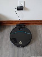 Robot vacuum unit on a wood floor sitting in front of a charging dock plugged into a wall outlet.