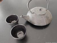 Teapot and two teacups on a black table showing design and condition.