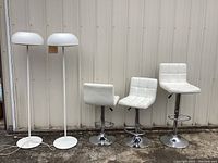Two modern white floor lamps with dome-shaped shades and circular bases alongside three white vinyl barstools with chrome bases placed outdoors near a metal wall.