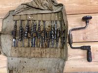 Full view of the antique hand brace drill next to canvas roll with drill bits inserted in pockets