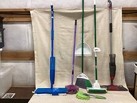 Full view of all six cleaning tools lined up including mops, broom, duster, dustpan and cobweb duster against cloth background.