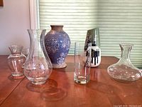 Seven vases and a glass decanter grouped on a wood table in front of a window with shade