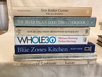 Shot of stacked cookbooks with titles visible including New Kosher Cuisine, The Silver Palate Good Times Cookbook, Whole30, The Blue Zones Kitchen, and Moosewood Cookbook.