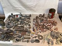 Wide view of assortment of metal plumbing parts including old faucets, various pipe fittings, clips, cut off pipe pieces, and brass casting waste spread out on brown paper.