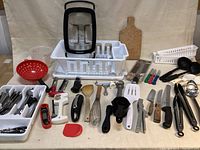 Photo showing a variety of kitchen tools and utensils laid out including dish racks, cutting board, strainer, spatulas, knives, and silverware divider with utensils.