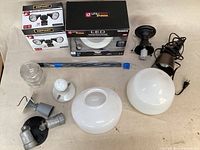 Assortment of light fixtures laid out together showing two boxed new lights, several used fixtures, two glass schoolhouse lamp shades, and extension rods.