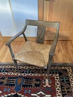 Front view of mid-century modern wood chair with woven seat and armrests on a patterned rug.
