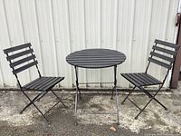 Front view of the metal folding patio table and two matching chairs set up side by side against a metal wall.