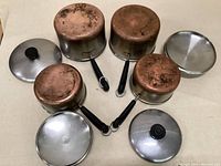 Four copper-bottomed pots with matching stainless steel lids arranged upside down showing the worn and tarnished copper bottoms and black handles.