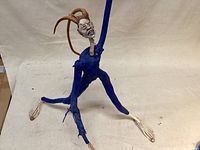 Close-up of humanoid sculpture with blue limbs, clay detailed face and bone-like hands on floor.