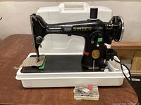 Singer black metal sewing machine atop white plastic carrying case with accessories box in front.