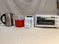 Front view of four appliances: stainless electric kettle, red rice cooker, white Cuisinart toaster, and white Black & Decker toaster oven.