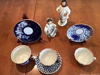 Overall view of lot including two Yakut girl figurines, three blue hand painted Lomonosov teacups with matching saucers, and two wooden display holders