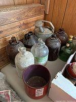 Eight glass jug bottles arranged on a wooden surface, a small red plant pot in front, and a metal watering can behind the bottles.