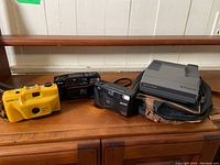 Four vintage film cameras on wooden surface including yellow Ultronic, black Ricoh, black Braun-Bravo, and gray Polaroid Spectra System with case.