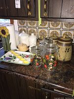 Various kitchen canisters including glass jars with floral designs, ceramic canisters, a sunflower-themed vase or tray, and egg-shaped yellow ceramic dish on a dark marble countertop.