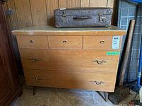 Front view of the cedar chest showing the faux-drawer front, three small upper drawers with round knobs, two large drawers with decorative handles, and faux-stone paper top. An old brown suitcase is placed on top of the chest.