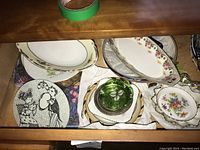 Photo showing a drawer with a variety of decorative plates and dishes, some stacked, others visible with floral and geometric patterns and a green glass piece.