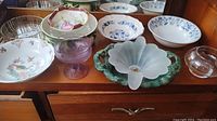 Wide view of the lot on wooden furniture showing multiple ceramic and glass bowls and dishes including floral patterns and a shell-shaped dish.