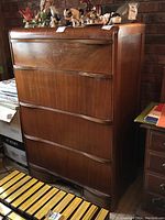 Frontal angled photo showing four drawer wood dresser with wear on drawer edges, medium brown finish, decorative items on top, and surrounding room context.