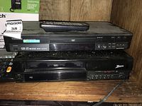 Toshiba DVD player stacked on Zenith VHS player on wooden shelf, dusty with visible wear.