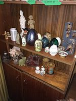 Shelf showing variety of figurines including ceramic woman, child, small angel figurines, decorative glass eggs in different colors including green and floral painted, a large conch shell, and various small decorative items including a red glass leaf and small ceramic birds