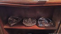 Three glass ashtrays on a wooden shelf: two with swirled designs and one cut glass ashtray in center showing detailed patterns.