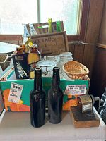 Photo of various vintage green and brown glass bottles along with clear jars and a small wicker basket placed inside orange and brown boxes.