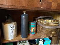 Shelf with two-tone crockery jug, tall dark bottle, colorful cooking powder tin, and large picnic tin with wooden handles.