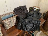 Two black leather duffle bags and one folding cloth sewing bag on a wooden surface near other household items
