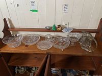 Shelf with multiple crystal bowls, two glass serving dishes with metal handles, mantle clock, and small animal figurines