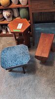 Photo showing three wooden furniture items on a carpeted floor: a small hexagonal side table, a solid wood stool with blue fabric top, and a solid wood bench-style stool.