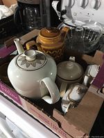 Multiple ceramic pots and porcelain containers including white teapot, brown coffee pot, and sugar and creamers in a cardboard box