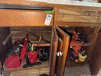 Wooden cabinet with open doors revealing red metal toolbox filled with hand tools, extension cords on shelf, window squeegee, trouble light, and other miscellaneous items.