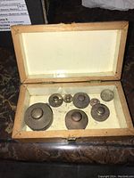 Closed wooden box containing an assortment of seven metal weights with knob handles, lined inside.