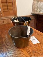 Photo showing two brass trade pails on a wooden surface, each with metal handles and showing wear consistent with age.