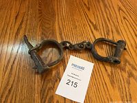 Photo showing the full antique handcuffs with chain and locking key, placed on wood surface with lot tag.