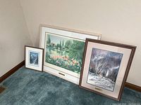 Three framed art prints placed on carpeting against a wall. Large floral landscape in white frame, medium winter lighthouse scene in brown frame, small mountain scene in white frame.