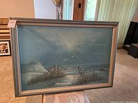 Framed coastal seascape painting with visible beach grass, fence posts, seagulls, and ocean scene.