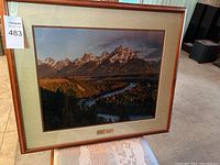 Large framed photograph showing a mountain range with forest and river in foreground, wood frame.
